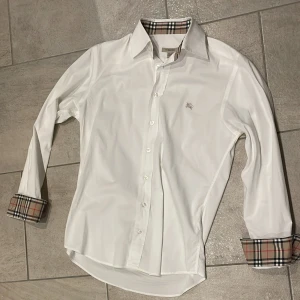 Vit Burberry skjorta med rutiga detaljer - Vit skjorta från Burberry med klassiska rutiga detaljer på insidan av kragen och manschetterna. Skjortan har lång ärm, knappar framtill och en liten broderad Burberry-logga på bröstet. Snygg och stilren modell i bomull.
