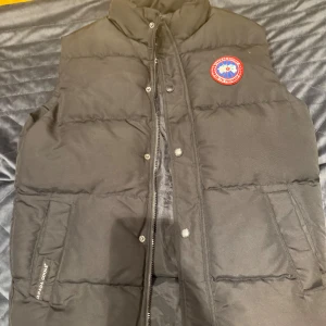 Svart dunväst från Canada Goose S - Svart dunväst från Canada Goose med klassisk logga på bröstet. Västen har hög krage, dragkedja och tryckknappar framtill samt två stora fickor. Fylld med vitt ankdun och polyester. Perfekt för kalla dagar när du vill hålla stilen.