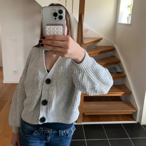 Ljusgrå chunky stickad kofta från H&M - Supermysig ljusgrå stickad kofta från H&M med stora svarta knappar och ribbade muddar. Koftan är croppad och har en loose fit med långa ärmar. Perfekt att slänga över ett linne för en chill look.