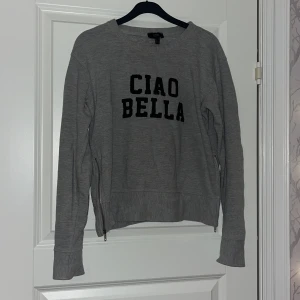 Grå sweatshirt från Mango med tryck - Grå sweatshirt från MNG Jeans med svart text 'CIAO BELLA' framtill. Tröjan har långa ärmar, ribbade muddar och coola dragkedjor i sidorna. 