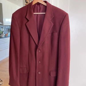 Vinröd kavaj från P & S Menswear - Säljer en vinröd kavaj från P & S Menswear i ull och kashmir. Kavajen har klassisk passform, tre knappar framtill, fickor med lock och en bröstficka. Tyget är tillverkat i Italien och har en mjuk, lyxig känsla. Perfekt för dig som vill sticka ut med stil.