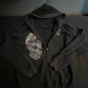 Svart hoodie från bvaldi - Svart hoodie med dragkedja och huva. På framsidan finns en stor strassdekor i form av en mask och på ryggen står det 'BANK ROBBER' i glittriga stenar. Hoodien har långa ärmar och en cool streetstil.