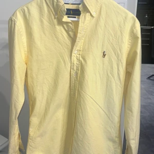 Gul slim fit skjorta från Ralph Lauren - Fräsch gul skjorta från Ralph Lauren i slim fit-modell. Klassisk button down-krage, broderad logga på bröstet och långa ärmar. Tillverkad i mjuk bomull och har knappar framtill. Perfekt för dig som gillar stilrena och färgglada plagg.