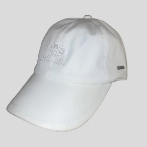 Vit keps från Loro Piana  - Exklusiv vit keps från Loro Piana Storm System med broderad logga framtill och metallmärke på sidan. Kepsen har en klassisk böjd skärm och insidan är fodrad med rutigt tyg i beige och bruna toner. Perfekt accessoar för en clean och stilren look.