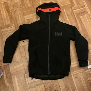 Svart vindjacka Helly Hansen med huva - Svart vindjacka från Helly Hansen med stor huva och orange detalj med HH-logga. Jackan har dragkedja framtill, två sidofickor med dragkedja och en bröstficka. Perfekt för blåsiga dagar och outdoor-aktiviteter. Snygg, sportig look med diskret HH-logga på bröstet. Köpte den för 4700kr säljer den för 2759