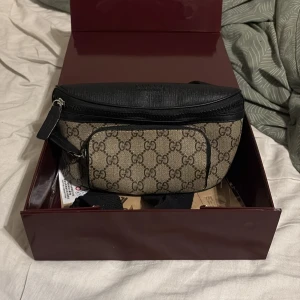 Gucci axelväska med GG-mönster - Snygg axelväska från Gucci i beige och svart med klassiskt GG-mönster. Väskan har svarta skinn- och textildetaljer, justerbar rem och flera dragkedjefack. Perfekt för dig som vill ha en stilren och lyxig accessoar med tydlig logga.