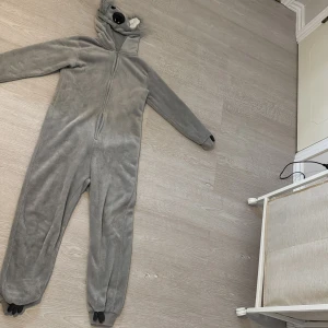 Grå fleece onesie med huva - Supermysig grå onesie i fleece med huva som har söta öron och broderat djuransikte från Lindex. Dragkedja framtill och långa ärmar samt ben med muddar. Perfekt för chilliga dagar hemma eller som rolig pyjamas.