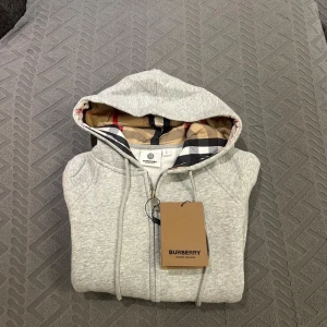 Burberry hoodie grå - Snygg grå hoodie från Burberry med klassiskt rutigt foder i huvan. Tröjan har dragkedja framtill, snörning i huvan och två fickor. Perfekt för dig som vill ha en stilren och lyxig look med ikoniska Burberry-detaljer. STORLEKEN ÄR S MEN KÄNNS SOM XS