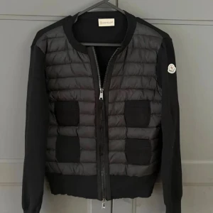 Svart cardigan jacka från Moncler - Jag säljer min Moncler cardigan 🖤. Den är svart med quiltad, lite glansig front, dragkedja fram och två söta fickor med knappar. Stickade ärmar och ribbade muddar gör den både mysig och stilren. Självklart med den klassiska Moncler-loggan på ärmen 🤍. Den är perfekt om du vill ha en lyxig men ändå vardagssnygg look ✨. Säljer bara för att jag har lite för många jackor 🙈.