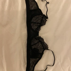 Svart spetsig bralette med tunna band - Säljer en svart bralette med tunna axelband och vacker spetsdetalj. Toppen har genomskinlig spets med blommönster, aldrig använt. Strl 75B men den är rätt liten skulle jag säga