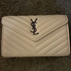 Vit quiltad axelväska från Saint Laurent - Snygg vit axelväska från Saint Laurent med klassisk YSL-logga i metall på framsidan. Väskan har quiltad design, kedjerem i mörk metall och flera fack inuti, inklusive ett med dragkedja. Perfekt storlek för det viktigaste och riktigt stilren look.  OPS INTE ÄKTA!
