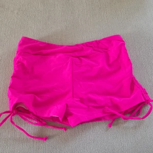 Neonrosa badshorts med snörning - Säljer ett par neonrosa badshorts med snörning i sidorna och rynkdetalj bak. Shortsen är tillverkade i ett stretchigt material av nylon och spandex, perfekta för stranden eller poolen. Supersnygga och bekväma med en riktigt poppig färg! Aldrig använda.