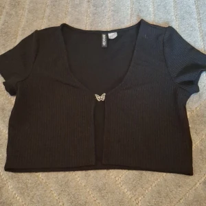 Svart croppad bolerotopp från H&M Divided - Svart ribbad bolerotopp från H&M Divided i croppad modell. Toppen har korta ärmar, djup v-ringning och en söt fjärilsdetalj framtill som stängning. Perfekt att styla över linne eller klänning för en trendig look.