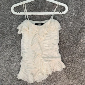 Vit volangig linnetopp från Bik Bok - Supersöt vit linnetopp från Bik Bok med tunna axelband och volangdetaljer framtill och nedtill. Toppen har ett strukturerat, veckat tyg som ger en unik look och är perfekt för varma dagar. Lätt och luftig känsla, passar snyggt till både jeans och kjol.