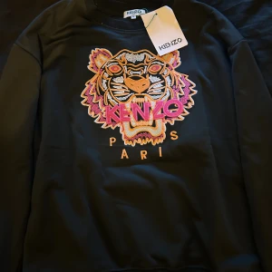 Svart Kenzo sweatshirt med tigerbrodyr - Svart sweatshirt från Kenzo med ikonisk broderad tiger i orange, rosa och gult framtill. Tröjan har rund halsringning, långa ärmar och ribbade muddar. Perfekt statement-plagg med färgstarka detaljer och klassisk Kenzo Paris-logga.