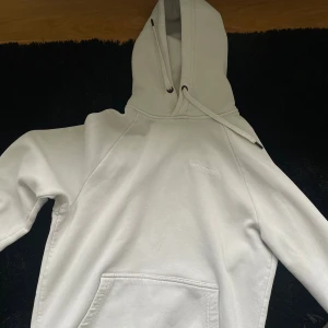 Vit hoodie från Peak Performance - Säljer en stilren vit hoodie från Peak Performance med broderad logga på bröstet. Tröjan har huva med snörning och en stor magficka. Perfekt för dig som gillar en clean och enkel look.