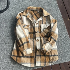 Rutig overshirt i beige och brunt - Overshirt med rutigt mönster i beige, brunt och svart. Jackan har klassisk krage, knäppning framtill med bruna knappar och två bröstfickor med lock. Tillverkad i mjuk polyester, perfekt för lager-på-lager under höst och vår.