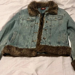 Jeansjacka med fuskpäls från Flash - Snygg ljusblå jeansjacka från Flash med bruna fuskpälsdetaljer på krage, ärmslut och nederkant. Jackan har knappar framtill, två bröstfickor med lock och två sidofickor. Insidan är quiltad och fodrad i svart för extra värme.