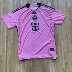Rosa Inter Miami fotbollströja Adidas - Säljer en rosa Inter Miami fotbollströja från Adidas med svarta detaljer och klubbmärke på bröstet. Tröjan har korta ärmar, svart logga och klassiska Adidas-ränder på axlarna. Tillverkad i lätt och ventilerande material, perfekt för match eller träning.