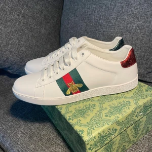 Gucci Ace vita sneakers med bi-detalj - Gucci Ace sneakers i vitt skinn med klassiska gröna och röda ränder på sidorna, broderad guldfärgad bi och metallicröd samt metallicgrön häl i ormskinnsmönster. Platt sula, snörning och rund tå. En ikonisk och stilren sneaker för dig som gillar lyxiga detaljer.
