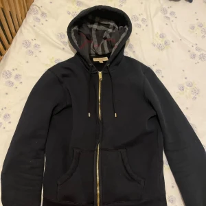  Burberry hoodie - Svart hoodie från Burberry med klassiskt rutigt foder i huvan och guldfärgad dragkedja. Tröjan har långa ärmar, känguruficka och justerbar huva med snörning. Perfekt för dig som vill ha en stilren och lyxig streetwear-look.
