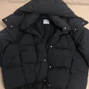 Svart pufferjacka bershka  - Säljer en svart pufferjacka från bershka med stor luva och knappar. Jackan är vadderad och har dragkedja samt tryckknappar framtill. Perfekt för kalla dagar och är oversized. 