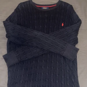 Mörkblå stickad tröja Polo Ralph Lauren - Kabelstickad mörkblå tröja från Polo Ralph Lauren med klassisk rund halsringning och röd broderad logga på bröstet. Långärmad modell i mjuk stickad bomull, perfekt för lager-på-lager under höst och vinter.