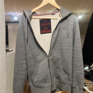 Grå hoodie med dragkedja från Superdry - Säljer en grå hoodie från Superdry med broderad logga på bröstet, huva med snörning och praktiska fickor framtill. Tröjan har hel dragkedja och är tillverkad i mjuk bomullsblandning. Perfekt för chill dagar eller när du vill ha en enkel streetstyle.