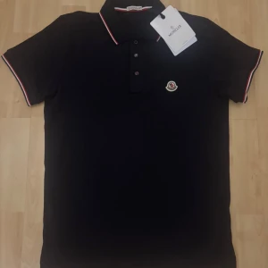 Svart pikétröja från Moncler - Snygg svart pikétröja från Moncler med klassisk krage och knäppning framtill. Tröjan har Moncler-logga på bröstet och röda samt vita detaljer på krage och ärmslut. Perfekt för dig som gillar stilrena och exklusiva plagg.