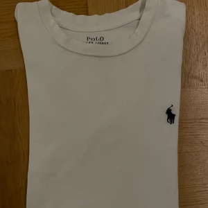 Vit tröja  från Polo Ralph Lauren - Vit tröja  från Polo Ralph Lauren med klassisk rund hals och broderad marinblå logga på bröstet. Tillverkad i mjuk bomull och har en clean, stilren look som funkar till allt. Perfekt för dig som gillar enkel och tidlös stil.