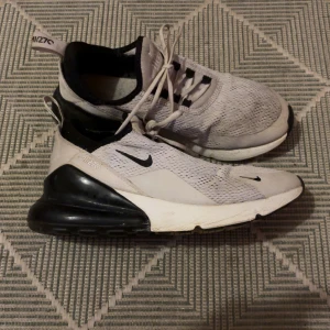 Nike Air Max 270 vita sneakers - Nike Air Max 270 sneakers i vitt med svarta detaljer och stor svart Air Max-häl. Ovandelen är i mesh för bra andningsförmåga och skon har snörning. Snygg och sportig design som passar perfekt till streetwear.