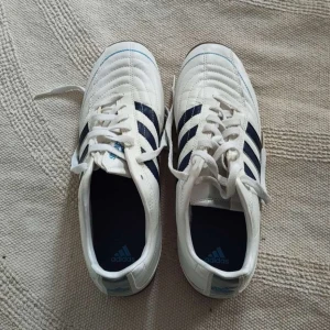 Adidas vita fotbollsskor strl 43 1/3 - Säljer ett par klassiska vita Adidas fotbollsskor med mörkblå ränder och bruna sulor. Skorna har snörning och är tillverkade i syntetmaterial med en stilren design. Perfekta för dig som vill ha snygga och bekväma skor till inomhusfotboll.