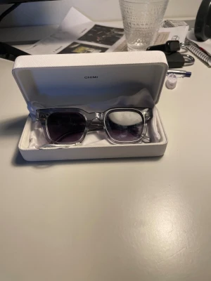 Chimi 04 - Säljer ett par snygga solglasögon från Chimi som är använda max 5 gånger under en sommar. Glasen är mörklila och bågarna har en cool glittrig detalj på sidorna. Kommer med originalfodral. Perfekt accessoar för att lyfta din stil.