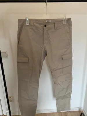 Beige cargobyxor från Dobber, W34 - Snygga beige cargobyxor från Dobber med raka ben och praktiska fickor på sidorna. Tillverkade i bomull, perfekt för dig som gillar en avslappnad och funktionell stil. Klassisk design med knapp och dragkedja framtill samt bälteshällor.