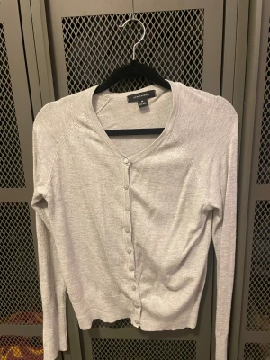 Ljusgrå kofta från Atmosphere - Ljusgrå kofta från Atmosphere i mjuk bomullsblandning. Klassisk v-ringad modell med långa ärmar och knappar hela vägen framtill. Perfekt att slänga över en t-shirt eller linne.