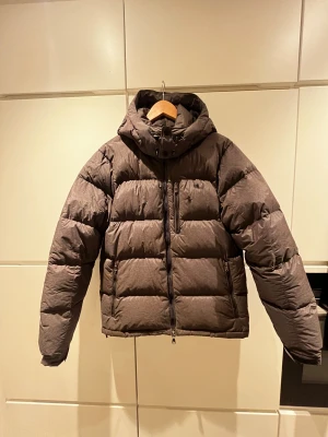 Grå dunjacka från Polo Ralph Lauren - Snygg grå dunjacka från Polo Ralph Lauren med broderad logga på bröstet. Jackan har huva, flera dragkedjeförsedda fickor och en rejäl dragkedja framtill. Perfekt för kalla dagar och har en klassisk pufferstil som är både trendig och bekväm. OBS brännskada på vänster insida, är dock inget som syns eller märks. Storleken är M/L.