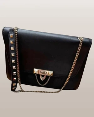 Svart Valentino axelväska med nitar - Snygg svart axelväska från Valentino i skinn med guldfärgade detaljer och kedja. Väskan har rektangulär form, dekorativa nitar på axelremmen och ett elegant spänne framtill. Insidan har dragkedjefack för småsaker. Perfekt för dig som gillar lyxiga accessoarer.