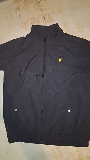 Svart vindjacka från Lyle & Scott - Svart vindjacka från Lyle & Scott med klassisk gul logga på bröstet. Jackan har hel dragkedja framtill, två fickor med tryckknappar och hög krage. Tillverkad i lätt polyester, perfekt för blåsiga dagar. Enkel och stilren design som funkar till det mesta.