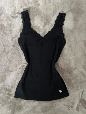Svart spetslinne Abercrombie & Fitch - Svart ribbad linnetopp från Abercrombie & Fitch med breda axelband och snygga spetsdetaljer längs v-ringningen. Liten vit logga nertill. Perfekt att styla med jeans eller kjol för en chill och trendig look. Vintage storlek M passar xs-m