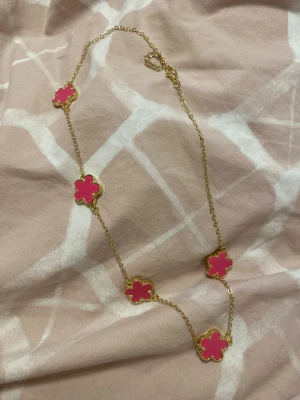 Guldfärgat halsband med rosa blommor - Superfint halsband med tunna guldfärgade kedjor och fem rosa blommor i emalj. Blommorna har en söt, rundad form och är jämnt placerade längs kedjan. Perfekt för dig som vill ha en färgklick och lekfull detalj i din stil.