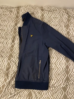 Blå jacka från Lyle & Scott XXL - Snygg mörkblå bomberjacka från Lyle & Scott med gul logga på bröstet. Jackan har ribbade muddar, två fickor med dragkedja och hel dragkedja framtill. Tillverkad i polyester och har en klassisk, stilren look som passar perfekt till streetwear. Passar mig som  L perfekt 