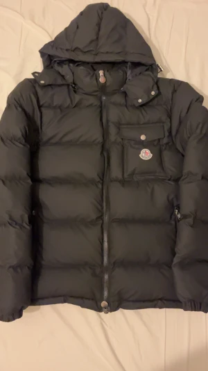 Svart Moncler pufferjacka med huva - Svart pufferjacka från Moncler i storlek S. Jackan har avtagbar huva, dragkedja framtill, bröstficka med logga och två sidofickor med dragkedja. Klassisk dunjacka med snygg, puffig look och diskreta detaljer i silver.