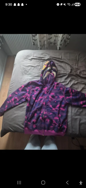 BAPE Shark Hoodie lila/camo - Säljer en ikonisk BAPE Shark Hoodie med lila och rosa camouflage-mönster. Hoodien har broderade detaljer med hajansikte och WGM-bokstäver på huvan, samt dragkedja hela vägen upp. Grym streetwear-vibe och riktigt unik design. Den är XXL men passar som en L