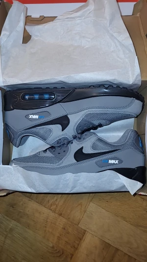 Nike Air Max 90 grå/blå sneakers - Nike Air Max 90 sneakers i grått med svarta detaljer och blå logga. Fick de i present men de passar tyvärr inte mig. HELT OANVÄNDA. Ovandel i mesh och syntet, svart sula med synlig Air Max-enhet. Klassisk siluett med snörning och rund tå. Perfekta för dig som gillar stilrena och sportiga vibes.