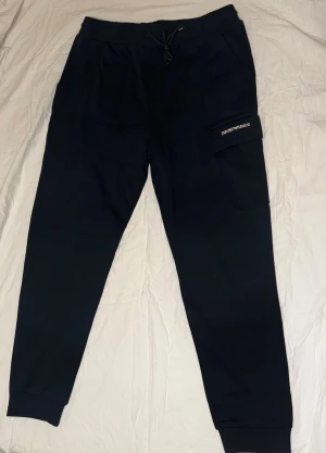 Svarta mjukisbyxor Emporio Armani - Svarta mjukisbyxor från Emporio Armani med snörning i midjan och en stor ficka med logotyp på benet. Byxorna har ribbade muddar vid bensluten och en relaxed passform som är både chill och snygg. Perfekt för en casual look.