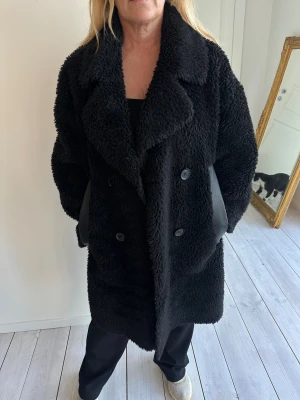 Svart teddyjacka med stora slag. - Mysig svart teddyjacka med oversized passform och stora slag. Jackan är knälång, har dubbelknäppning med knappar och sidofickor med detaljer i blankt tyg. Perfekt för kalla dagar när du vill vara både varm och snygg.