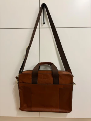 Brun datorväska i skinn från Saddler - Stilren brun datorväska från Saddler i äkta skinn med justerbar axelrem i textil och dubbla handtag. Väskan har flera fack på utsidan och detaljer i mässingsfärgad metall. Perfekt för att bära laptop och viktiga prylar till plugget eller jobbet.