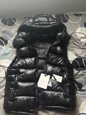 Svart dunväst från Moncler med huva - Svart glansig dunväst från Moncler med avtagbar huva, två dragkedjefickor och klassisk logga på bröstet. Västen har quiltad design och dubbla dragkedjor framtill. Perfekt för lager-på-lager och streetstyle.