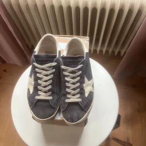 Golden Goose sneakers i mocka - Snygga Golden Goose sneakers i mörkblå mocka med vita detaljer och ikonisk stjärna på sidan. Skorna har vita snören, rund tå och platt sula. Bakpartiet och stjärnan är i vitt skinn, vilket ger en schysst kontrast till den mörka ovandelen.
