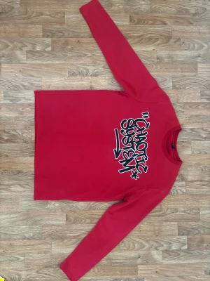 Röd långärmad tröja FSBN Chaotic System - Röd långärmad tröja från FSBN i storlek L med trycket 'Chaotic System' i svart och vitt på framsidan. Tröjan har en klassisk rund hals och är tillverkad i mjuk bomull. Regular fit för en avslappnad look.